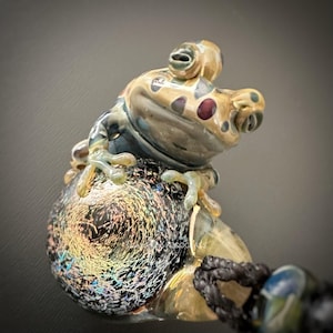 Puede incluir: Una detallada escultura de rana de vidrio con una base iridiscente. La rana es de color beige, azul y marrón con manchas rojas. La escultura está unida a un cordón negro.