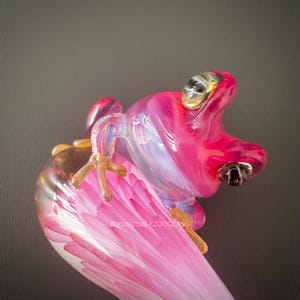 Puede incluir: Escultura de rana de cristal en tonos rosa, blanco y dorado. La rana tiene ojos grandes y detallados y patas delicadas. La escultura está posada sobre una estructura en forma de pétalo rosa y blanco. El texto "©2021-2026 KZOOGLASS" es visible.
