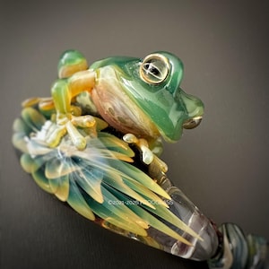 Puede incluir: Una detallada escultura de rana de cristal en tonos verdes, amarillos y transparentes. La rana está posada sobre una estructura texturizada en forma de hoja con un degradado de colores. La escultura tiene detalles intrincados, incluyendo los ojos y las patas de la rana. El texto "©2021-2026 WOODGLASS" es visible.