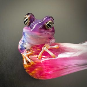 Puede incluir: Una escultura de rana de cristal en tonos morados, rosas y naranjas. La rana está posada sobre un pétalo rosa y naranja, con detalles y un acabado brillante. Una pieza de arte única.