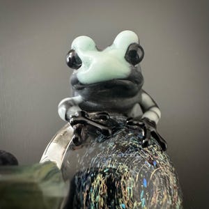 Peut inclure: Figurine de grenouille en verre avec une tête vert clair et des yeux noirs, assise sur une base texturée. Le corps de la grenouille est rayé noir et blanc. La base a un design tacheté coloré. La figurine est un objet décoratif unique.