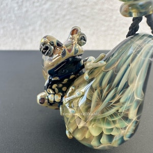 Puede incluir: Un colgante de cristal detallado con forma de rana en tonos verdes, azules y dorados. La rana está posada sobre una pieza de cristal en forma de lágrima con un diseño de plumas. El colgante está sujeto a un cordón negro.