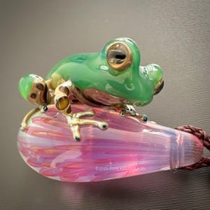 Puede incluir: Colgante de cristal con forma de rana verde y dorada, posado sobre una lágrima de cristal rosa y morada. El colgante tiene un cordón marrón y el texto "©2021-2025 KZOOGLASS".