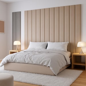 May include: A modern bedroom setup featuring a beige upholstered headboard with vertical panels. The bed is made with white bedding and pillows. Two small wooden nightstands with lamps flank the bed. A white rug and a small ottoman complete the scene.