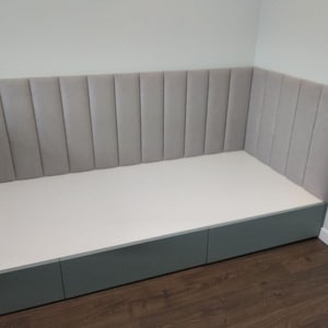 May include: A custom-built, corner bench seat with a light gray, vertically-paneled headboard. The base is a matching gray with three drawers. The seat is white.