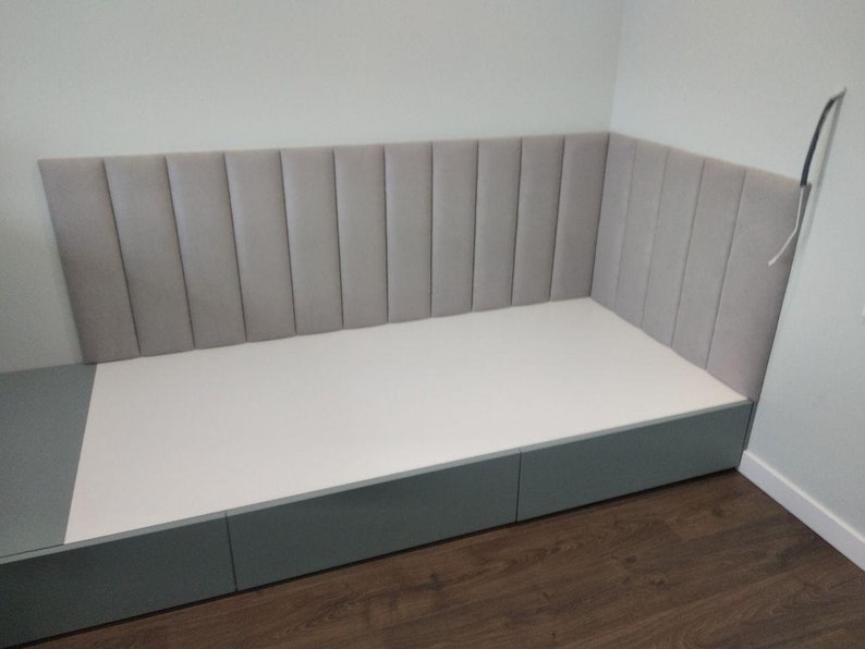 May include: A custom-built, corner bench seat with a light gray, vertically-paneled headboard. The base is a matching gray with three drawers. The seat is white.