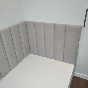 May include: A light gray upholstered headboard with vertical paneling, installed in a corner. The headboard is attached to a white bed frame. The wall is painted a light gray color.