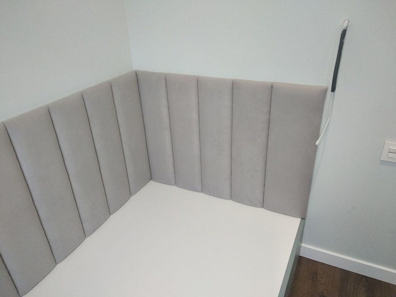 May include: A light gray upholstered headboard with vertical paneling, installed in a corner. The headboard is attached to a white bed frame. The wall is painted a light gray color.