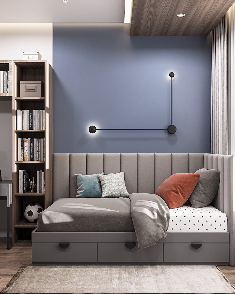 May include: A modern bedroom interior featuring a gray bed with a tufted headboard and three storage drawers. The bed is adorned with pillows in shades of blue, orange, and gray. A bookshelf and a soccer ball are visible. The wall is painted blue.