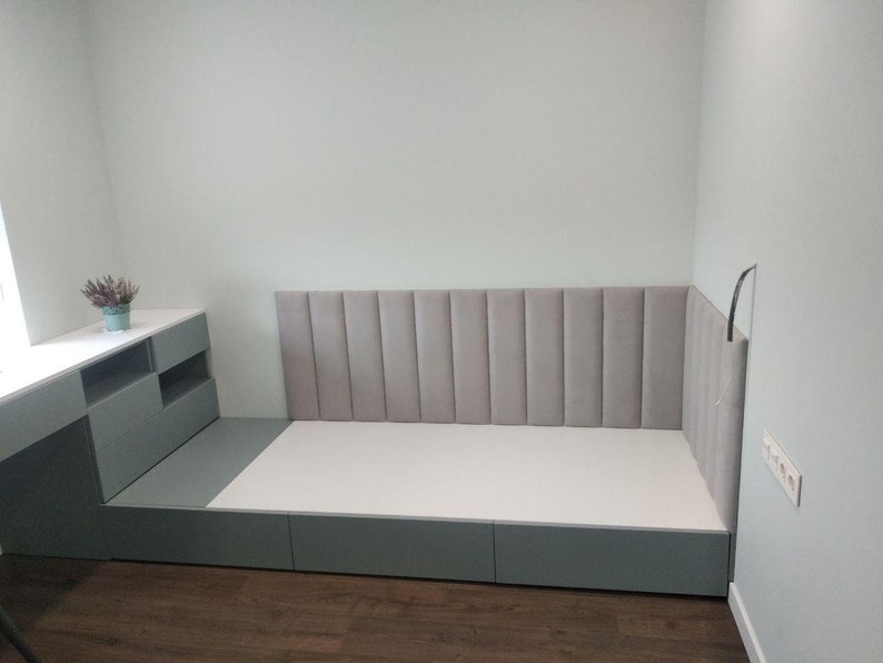 May include: A modern bed frame with a gray headboard and a white mattress. The bed is built into a corner, with a matching gray storage unit and desk. The headboard has vertical, padded panels.