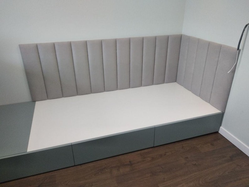 May include: A built-in bed frame with a gray upholstered headboard and a white base. The bed frame has a gray base with three drawers. The headboard is divided into vertical padded sections.