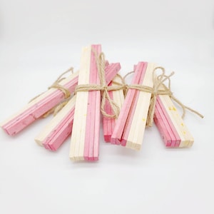 May include: Several bundles of craft sticks, each featuring a pink stripe, are secured with twine. The sticks are arranged on a white backdrop, suitable for crafting or decorative use. Each bundle is approximately 15 cm long.