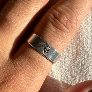 May include: A silver-colored ring with a bow design etched on the band. The ring is worn on a finger, and the bow is centered on the front of the ring. The ring has a flat, wide band.