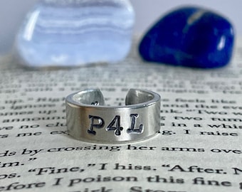 Könnte beinhalten: Ein verstellbarer Silberring mit den Buchstaben "P4L" auf der Vorderseite. Der Ring liegt auf einer Buchseite, mit einem blauen und einem hellblauen Stein im Hintergrund.