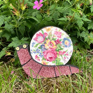 May include: A decorative snail sculpture featuring a body covered in a pink and black maze-like pattern with two yellow eyes. The snail shell is a circular piece of white ceramic decorated with a colourful floral print of pink, yellow, and blue flowers.