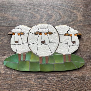 May include: A mosaic wall art piece featuring three white sheep with textured wool patterns, orange eyes, and brown ears. The sheep stand on a base composed of rectangular green glass tiles. The entire composition is constructed from individual pieces.