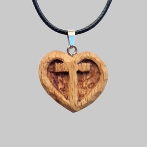 Può includere: Un ciondolo a forma di cuore in legno con un disegno a croce intagliata. Il ciondolo è appeso a una collana con cordino nero. Il legno ha una venatura naturale e un colore marrone caldo. Il ciondolo è sospeso da un'ansa color argento.
