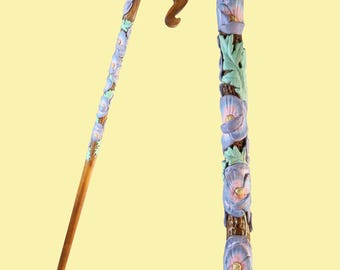 Puede incluir: Un bastón de madera con un diseño floral tallado. Las flores están pintadas en tonos de morado, rosa y verde. El bastón tiene un mango curvo.