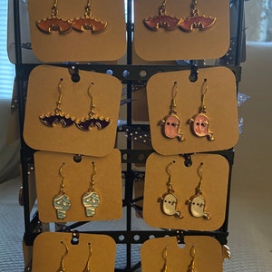 May include: A display of Halloween-themed earrings. The earrings are on a brown card with a black polka dot background. The earrings are gold with different coloured enamel designs. The designs include bats, ghosts, mummies, witches hats, and pumpkins.