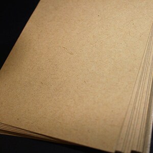 May include: A stack of blank, light brown paper sheets. The paper has a slightly textured surface and a natural, earthy tone. The edges of the paper are visible, showing the thickness of the stack. The background is black.
