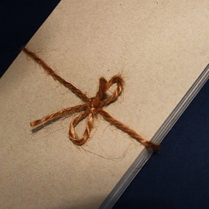 May include: A stack of beige paper tied with a brown twine bow. The paper has a slightly textured surface. The twine is knotted in a decorative bow. The background is a dark blue.