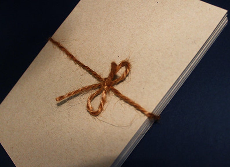 May include: A stack of beige paper tied with a brown twine bow. The paper has a slightly textured surface. The twine is knotted in a decorative bow. The background is a dark blue.