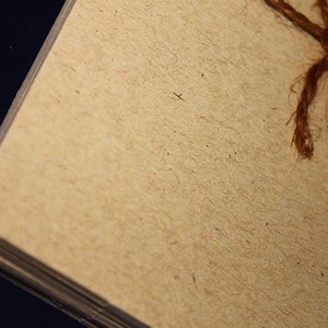 May include: Close-up of a stack of beige paper with rounded corners, bound with brown twine. The paper has a textured, fibrous appearance, and the twine is knotted at the top. The background is dark.