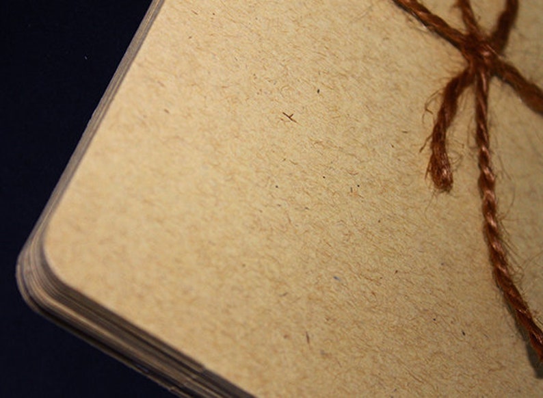 May include: Close-up of a stack of beige paper with rounded corners, bound with brown twine. The paper has a textured, fibrous appearance, and the twine is knotted at the top. The background is dark.