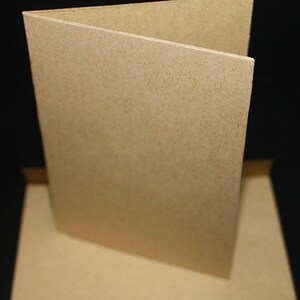 May include: A folded, blank greeting card made of light brown, textured paper. The card is open, revealing the inside, and sits on top of a matching envelope. The background is black.