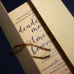May include: A kraft paper invitation with elegant calligraphy in dark blue ink. The invitation is tied with a brown twine and features event details, including names and the date. The paper has a natural, textured appearance.