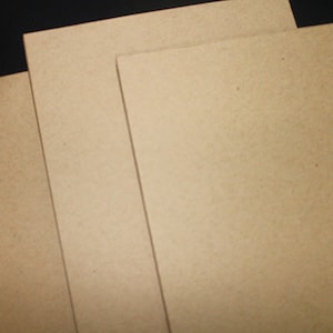 May include: A stack of several rectangular, light brown sheets of paper. The paper has a slightly textured surface and is arranged at an angle against a dark background. The sheets are likely for art or craft projects.