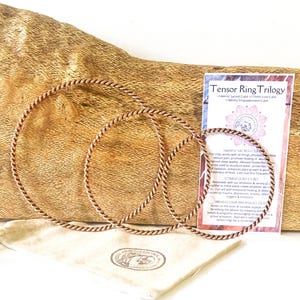 May include: Three copper-coloured metal rings with a twisted, rope-like texture, arranged in descending order of size. A rectangular information card titled Tensor Ring Trilogy and a small beige fabric pouch with a circular logo are included.