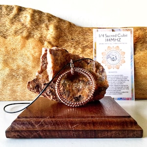 May include: A copper-coloured pendant with a circular woven design, suspended on a black cord. The pendant rests on a rough, textured stone, displayed on a wooden stand. A card with text reading "1/4 Sacred Cubit 144MHz" is visible.
