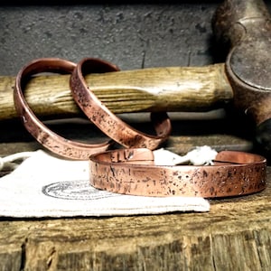 May include: Three copper bracelets with textured surfaces are displayed. Two are curved, and one is flat. A hammer and a canvas bag are in the background, suggesting a handcrafted jewellery theme.