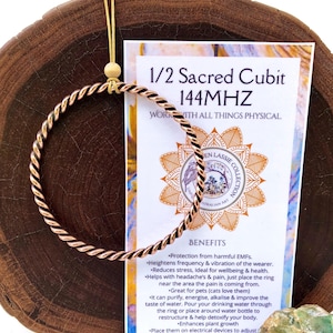 May include: A copper wire ring with a twisted design, labelled "1/2 Sacred Cubit 144MHZ" and "WORK WITH ALL THINGS PHYSICAL". The ring is on a wooden surface with a card describing its benefits, including protection from harmful EMFs, stress reduction, and energy purification.
