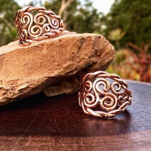 May include: Two copper rings featuring a swirling, Celtic knot design. The rings are displayed on a wooden surface.