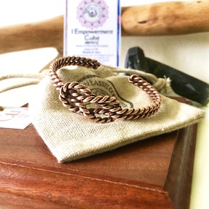 May include: A copper wire bracelet with a twisted knot design. The bracelet is displayed on a wooden surface with a small hessian pouch.