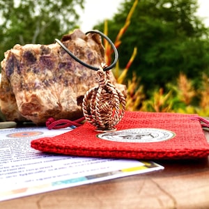 May include: A copper-coloured pendant necklace with a woven design, suspended on a black cord. The pendant sits on a red drawstring bag with a circular emblem. A large, rough-textured rock and printed materials are in the background.
