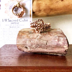 May include: A copper ring with a twisted design. The ring is on a piece of wood. The text "1/8 Sacred Cubit 54MHZ" is visible in the background.