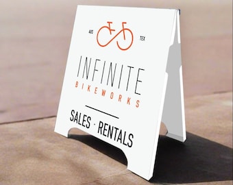 Puede incluir: Un cartel blanco en forma de A con el texto "Infinite Bikeworks" y "Sales - Rentals". El cartel también presenta un logotipo de bicicleta rojo con el texto "Aus Tex" encima.