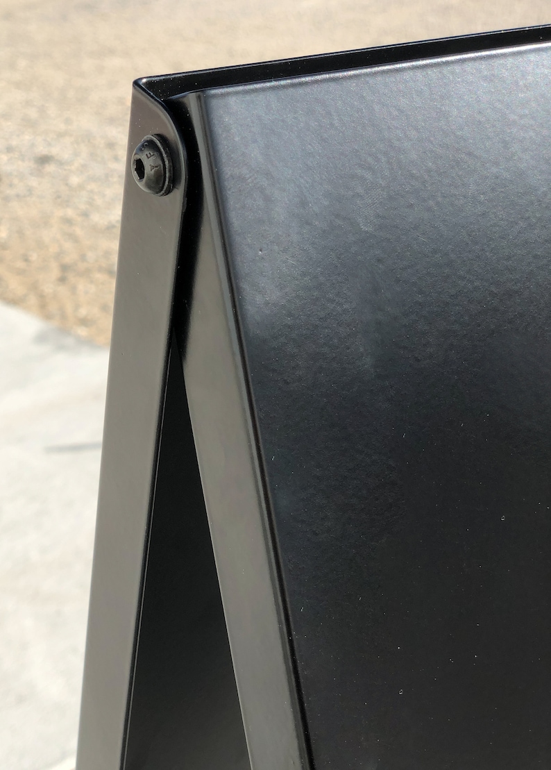 May include: Black metal A-frame sign with a black surface. The sign is made of two pieces of metal that are hinged together.