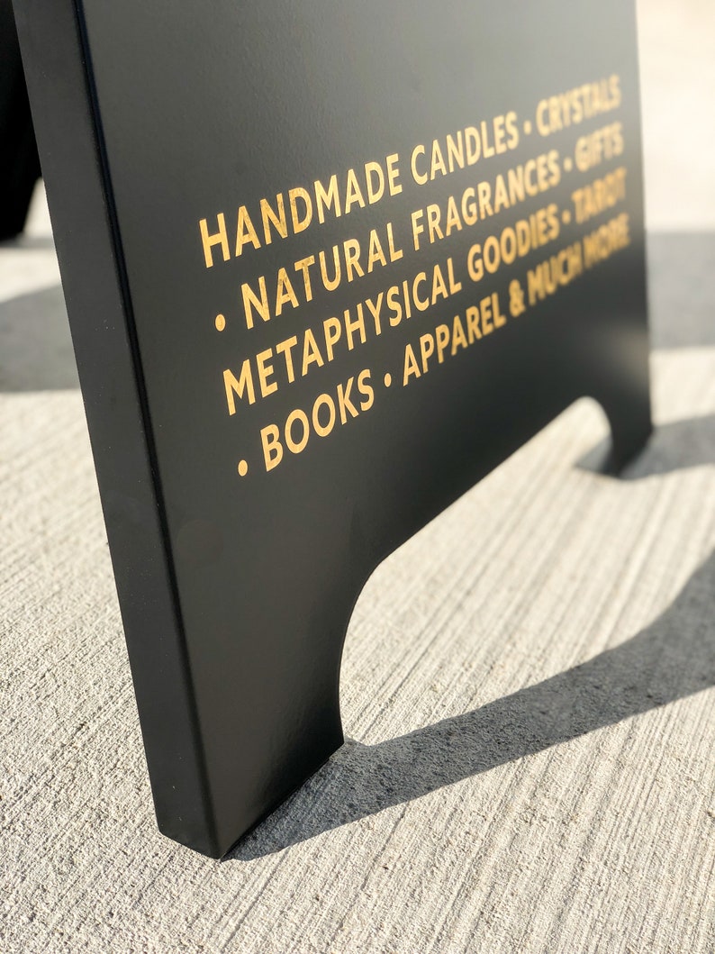 May include: A black A-frame sign with gold lettering advertising handmade candles, crystals, natural fragrances, gifts, metaphysical goodies, tarot, books, apparel, and much more.