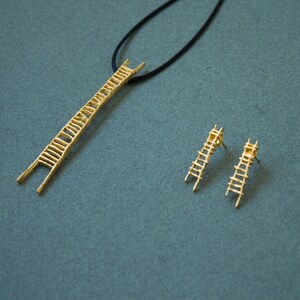 Pu&ograve; includere: Una collana d'oro con un ciondolo a forma di scala e orecchini a forma di scala abbinati. La collana &egrave; su un cordoncino nero.