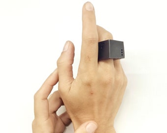 Può includere: Un anello quadrato nero con una superficie testurizzata. L'anello è indossato sull'indice di una mano.