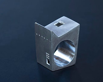 Può includere: Un anello color argento dal design geometrico. L'anello ha una forma rettangolare con una grande apertura circolare e un piccolo ritaglio quadrato. Presenta anche una serie di piccoli punti e linee distanziati.