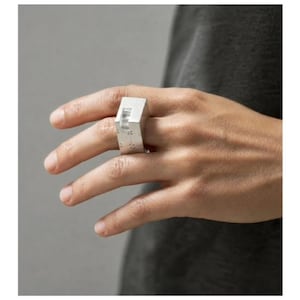 May include: A silver ring with a geometric design. The ring has a rectangular shape with a raised, angular element and small, evenly spaced dots. The ring is worn on a hand against a neutral background.
