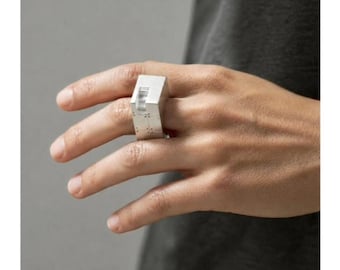 Può includere: Un anello color argento dal design geometrico. L'anello ha una forma rettangolare con un elemento angolare in rilievo e piccoli punti distanziati. L'anello è indossato su una mano su uno sfondo neutro.