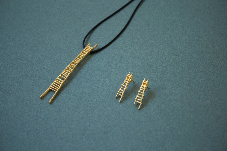 Pu&ograve; includere: Una collana d'oro con un ciondolo a forma di scala e orecchini a forma di scala abbinati. La collana &egrave; su un cordoncino nero.