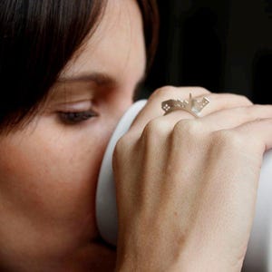May include: A close-up of a person's hand wearing a silver ring shaped like a building. The ring is on the index finger and the person is holding a white mug.