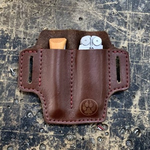 Puede incluir: Una funda doble de cuero marrón con costuras rojas. La funda tiene dos bolsillos, uno para un cuchillo pequeño y otro para una multiherramienta. La funda tiene un logotipo de huella de pata estampado en la parte delantera.
